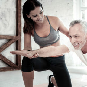 Motivated and strong. Radiant elderly gentleman smiling cheerfully while exercising with his fitness coach and training at a gym.